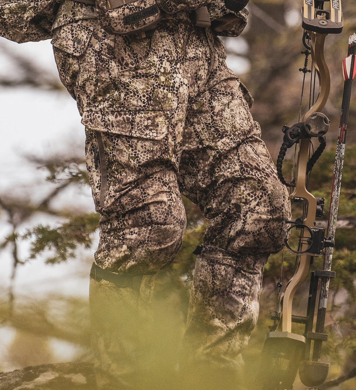 Hunter in Badlands camo pants, holding a compound bow; Badlands skull logo visible on gear.