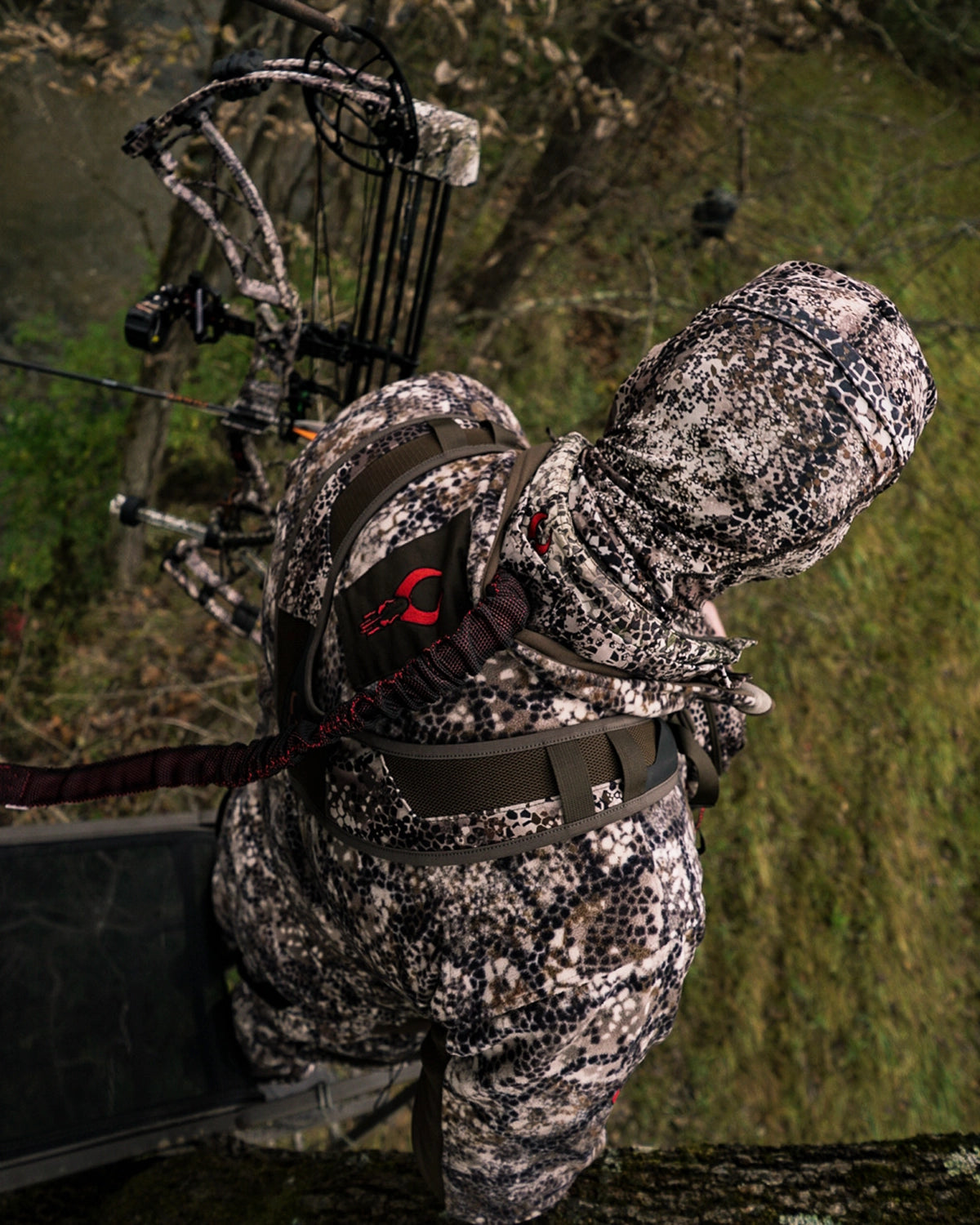 Hunter in camo gear with a bow, wearing a backpack featuring the red Badlands skull logo, stands on a tree stand.