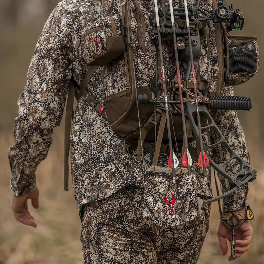 Hunter in Badlands camo with a bow and backpack; Badlands skull logo visible on gear, outdoors in a field.