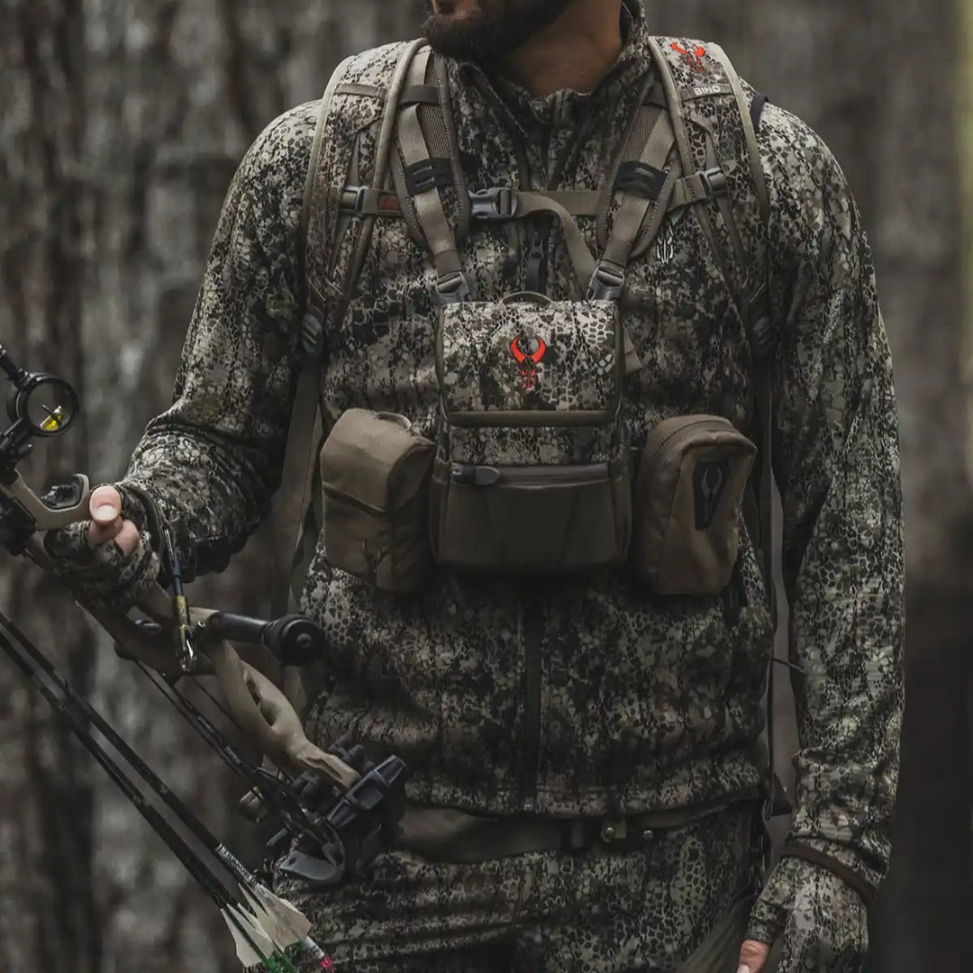 Hunter in Badlands camo gear, with Badlands skull logo on chest pack, holding a compound bow in a forest setting.