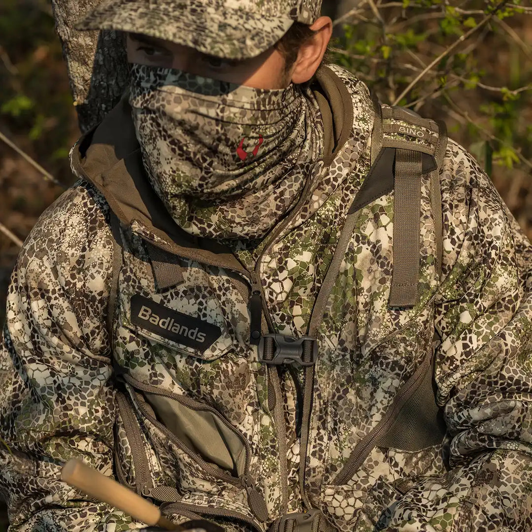 Person in Badlands camo gear outdoors; Badlands logo with skull visible on chest, surrounded by foliage.
