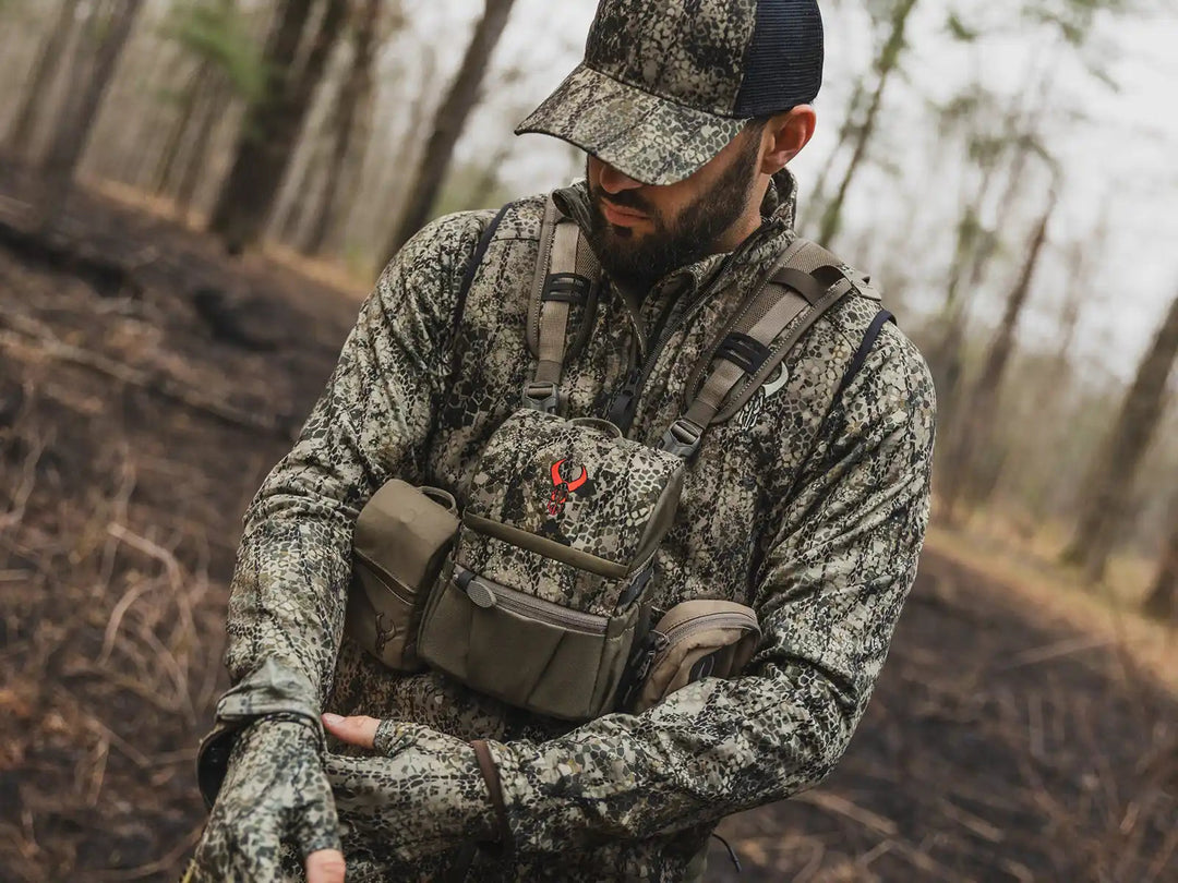 Person in camo gear and cap, wearing a Badlands chest pack with skull logo, standing in a forested area.