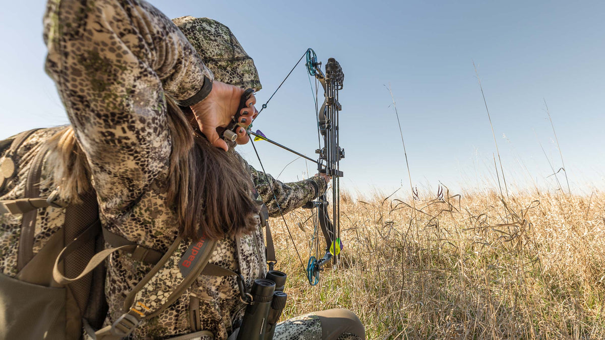 Hunter in Badlands camo draws a compound bow in a grassy field, Badlands skull logo visible on backpack strap.