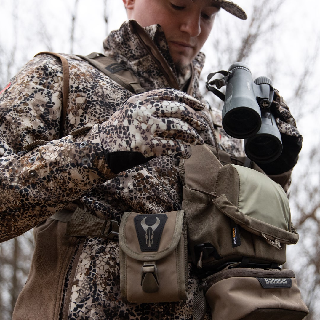 Person in Badlands camo gear holds binoculars; Badlands logo skull visible on pouch; outdoor woodland setting.