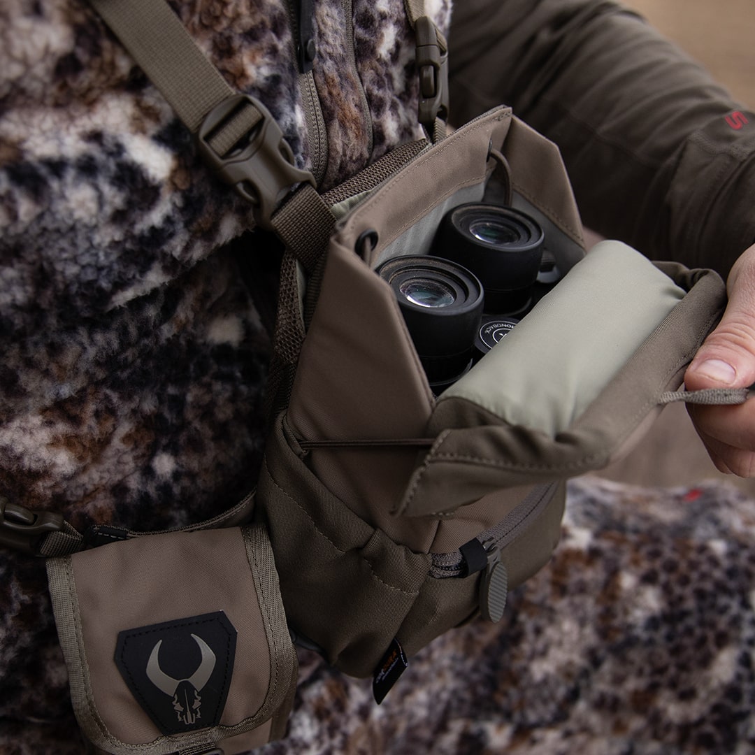 Person opens Badlands chest pack with logo, revealing binoculars inside; camo jacket visible in background.