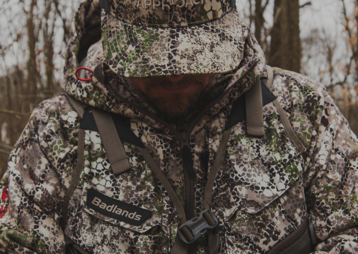 Hunter in Badlands camo jacket with Badlands logo and skull emblem, outdoors in a wooded area.