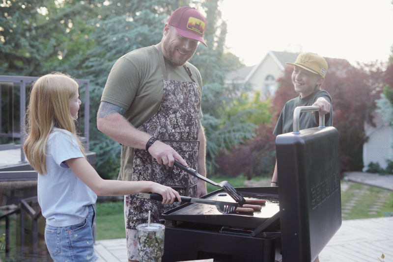 A man in a Badlands skull logo apron grills hot dogs outdoors with two children, surrounded by trees and a backyard setting.