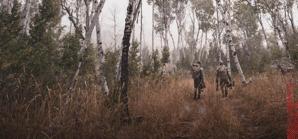 Two hunters in camo with Badlands skull logos walk through a misty forest of tall grass and aspen trees.
