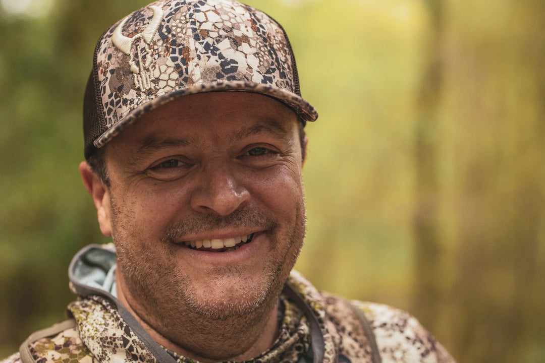Person wearing Badlands camo hat with skull logo and matching camo jacket, outdoors in a forest setting.