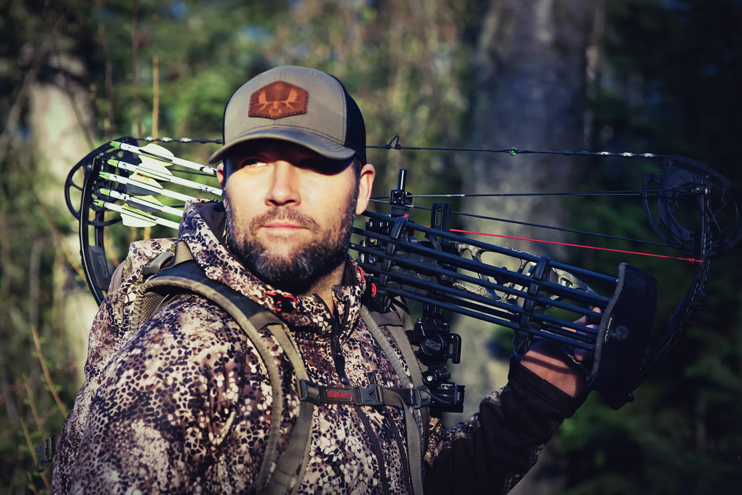 Hunter in Badlands camo with a compound bow and arrows, featuring the Badlands skull logo, standing in a forest.