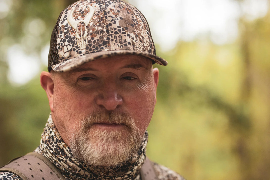 Person wearing Badlands camo hat with skull logo, matching neck gaiter, and backpack in an outdoor forest setting.