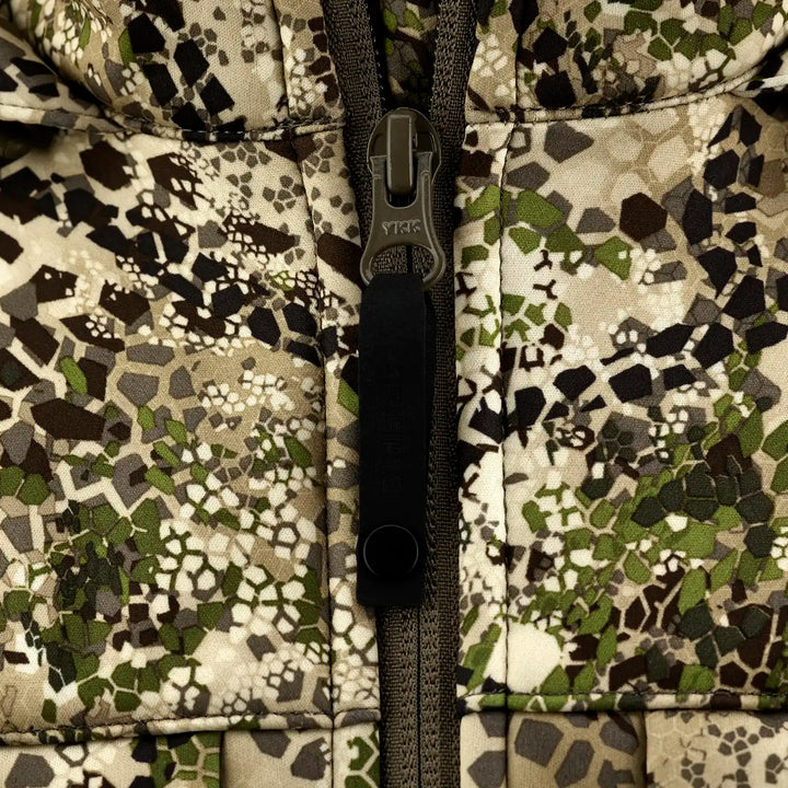 Close-up of Badlands camo jacket zipper; skull logo on pull tab, earth-tone hex pattern background.