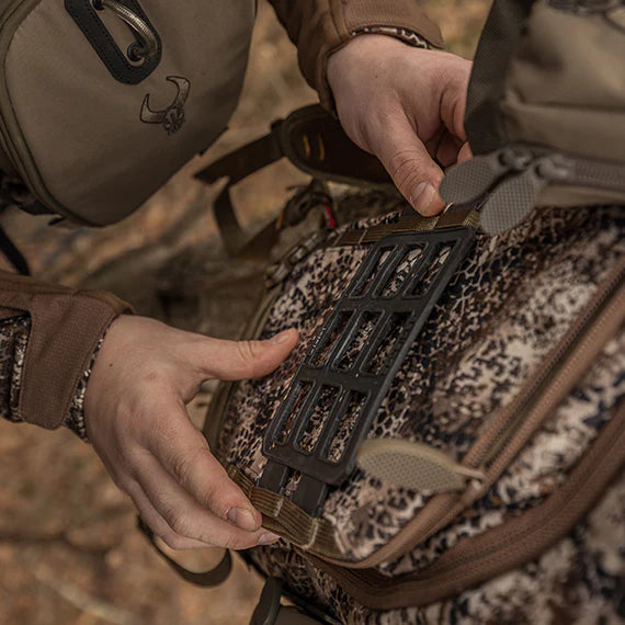 Hands attach gear to a Badlands camo backpack; Badlands skull logo visible on tan pouch.