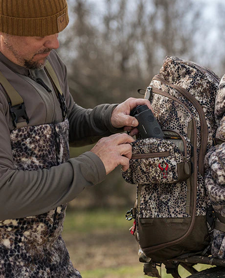 Person in camo gear places a device into a Badlands backpack with the skull logo, outdoors in a wooded area.