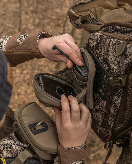 Person opens a Badlands camo backpack pocket; visible Badlands skull logo inside. Outdoor setting, hands in focus.