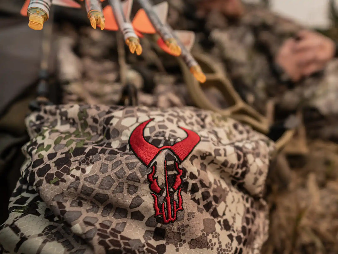 Close-up of a Badlands camo backpack with red skull logo, arrows above, and blurred hunter in background.