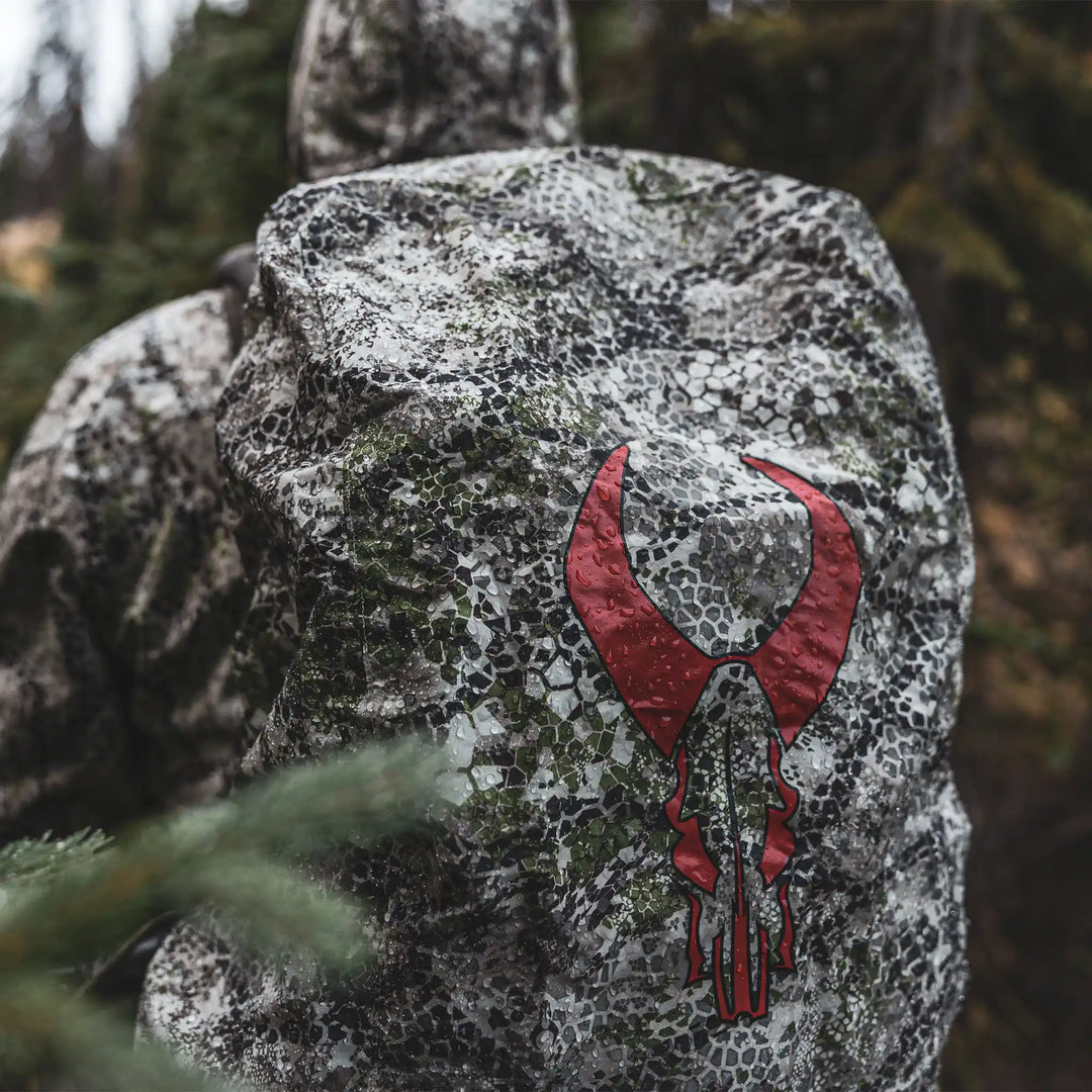 Hunter in camo gear with a Badlands backpack featuring a red skull logo, outdoors in a forest setting.