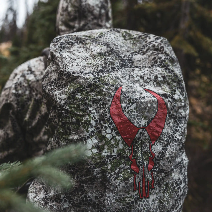 Hunter in camo gear with a Badlands backpack featuring a red skull logo, outdoors in a forest setting.