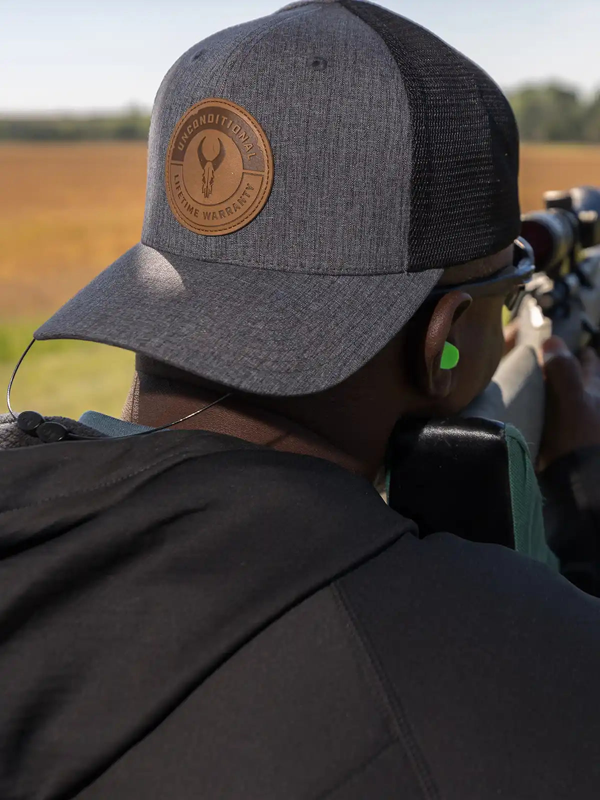 Person aiming a rifle outdoors, wearing a gray Badlands cap with skull logo and "Unconditional Lifetime Warranty" patch.