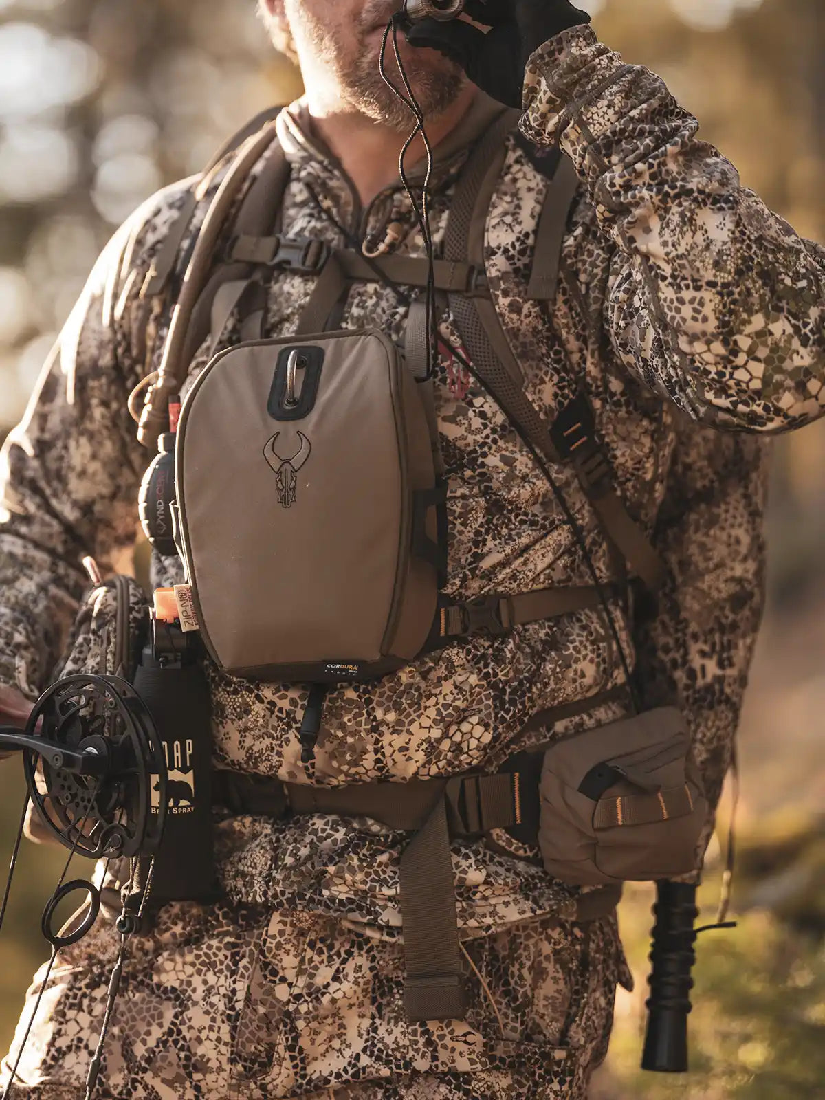 Hunter in Badlands camo gear drinks from a tube; chest pack features Badlands skull logo. Bow and bear spray visible.