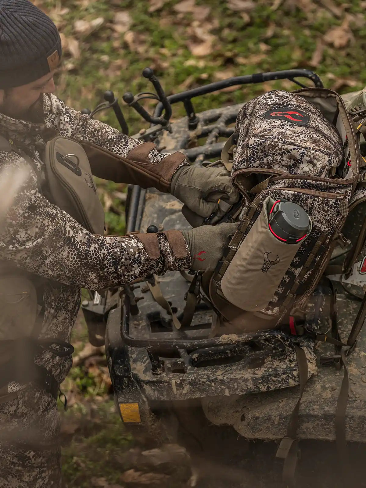 Hunter in camo gear loads a Badlands backpack with skull logo onto an ATV in a wooded area.