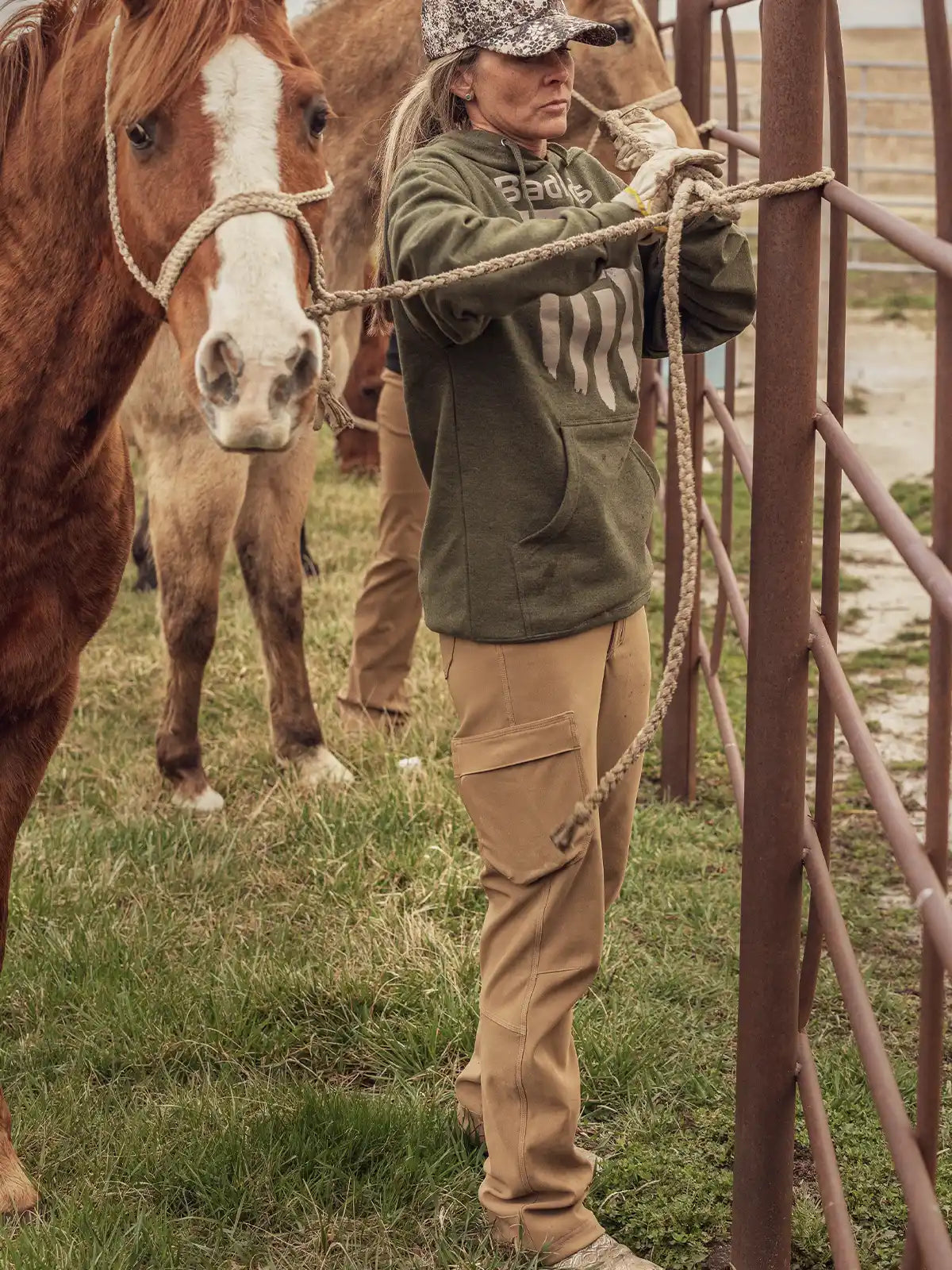 Person in Badlands hoodie with skull logo leads a horse near a metal fence on grassy ground, wearing tan cargo pants.