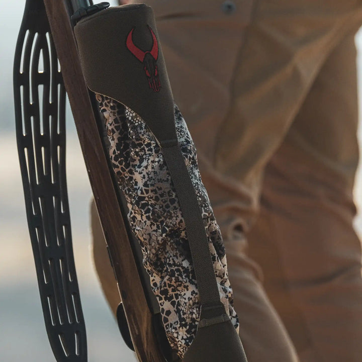 Close-up of a Badlands rifle sling with red skull logo and camo pattern, held by a person in tan outdoor pants.