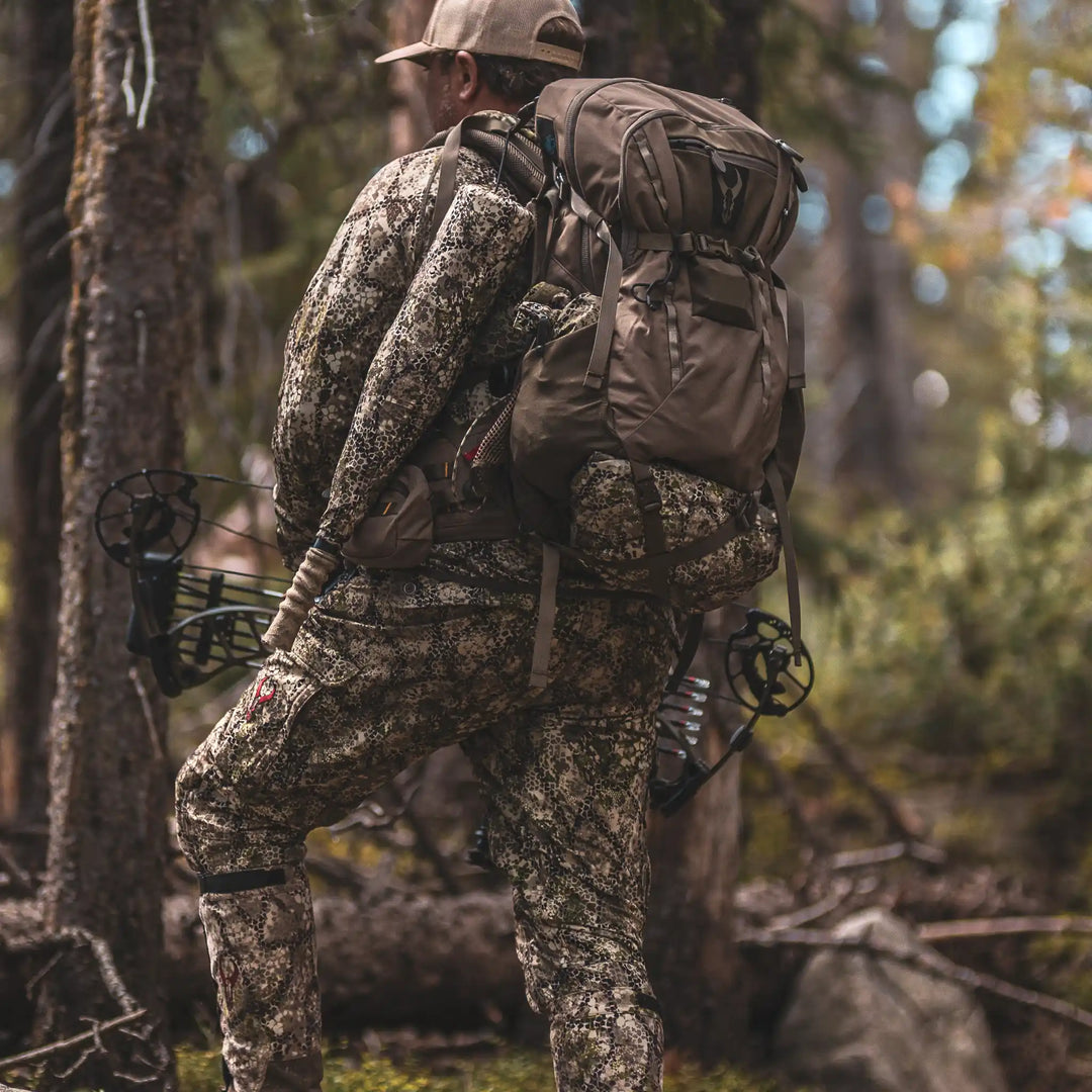 Hunter in camo gear with a Badlands backpack featuring the skull logo, carrying a bow in a forest setting.