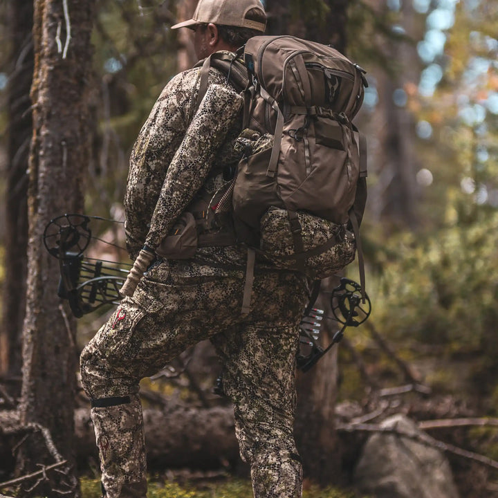 Hunter in camo gear with a Badlands backpack featuring the skull logo, carrying a bow in a forest setting.