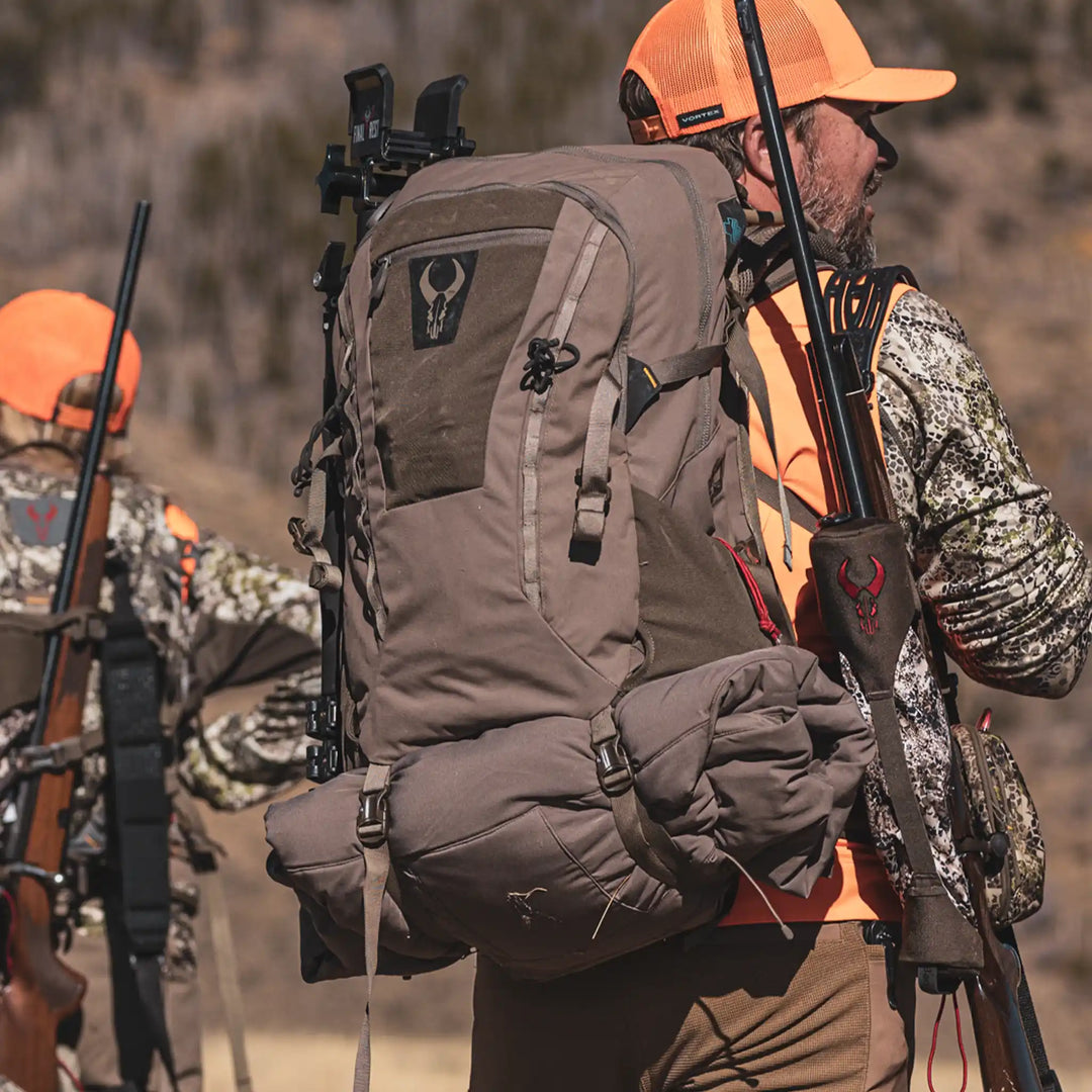 Hunters in camo and orange gear carry rifles and Badlands backpacks with the skull logo, outdoors in rugged terrain.