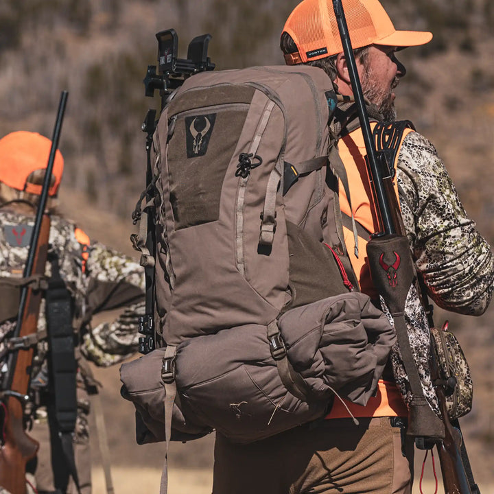 Hunters in camo and orange gear carry rifles and Badlands backpacks with the skull logo, outdoors in rugged terrain.
