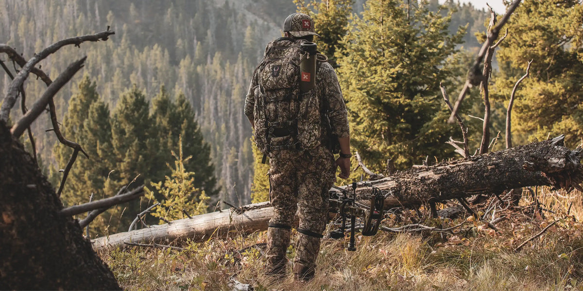 Hunter in camo gear with Badlands skull logo backpack stands in forest, holding a bow, facing pine trees and mountains.