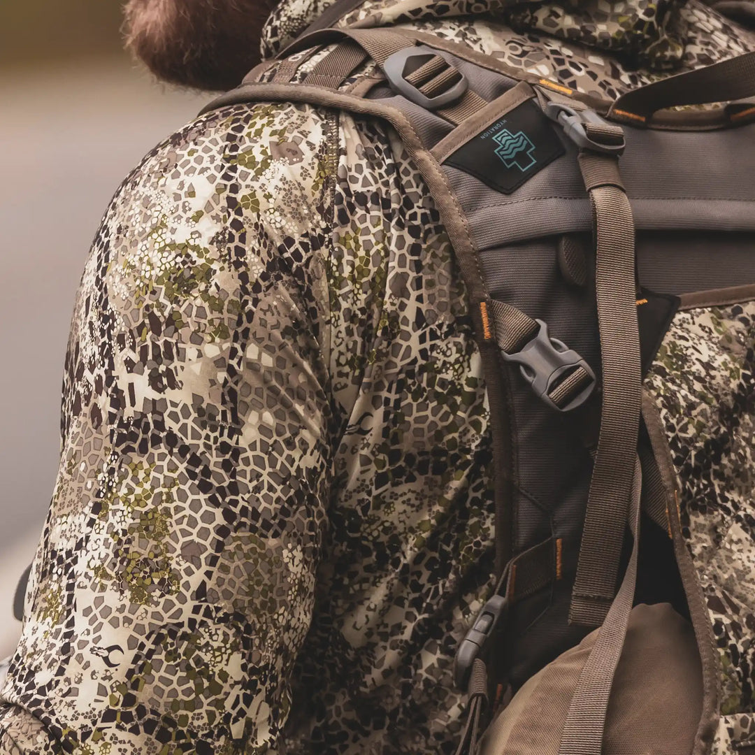 Person in camo jacket with Badlands skull logo on backpack strap; outdoor gear, close-up view.