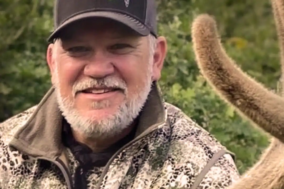 Person in Badlands camo jacket and hat with Badlands skull logo, outdoors near velvet antlers and green foliage.