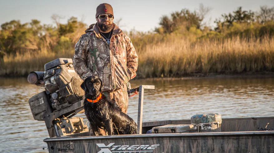 A hunter in Badlands camo stands on a boat with a black retriever, surrounded by marshland and calm water.