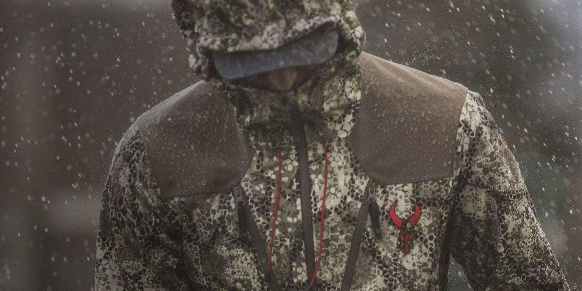 Person in camo rain jacket with Badlands skull logo, hood up, standing in falling rain.