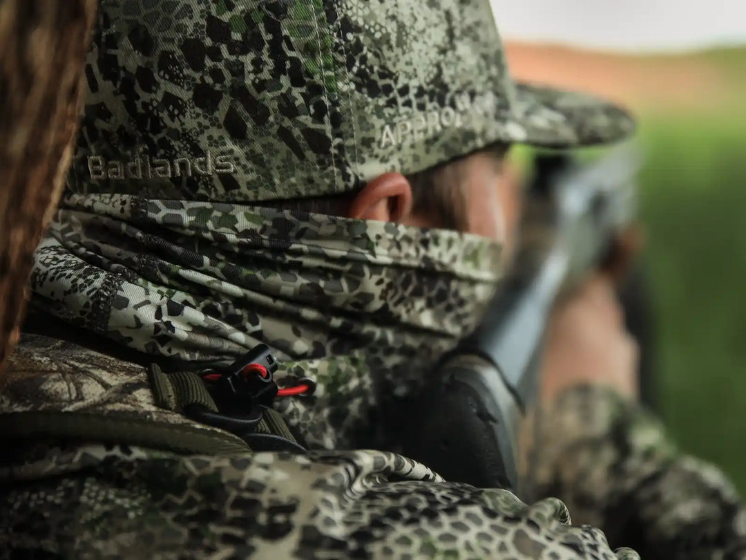 Hunter in Badlands camo gear aiming a rifle; Badlands logo visible on hat. Outdoor setting, focused on hunting.