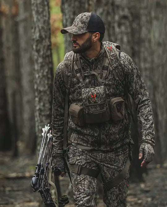 Hunter in Badlands camo gear with bow, wearing a pack featuring the red Badlands skull logo, walking through a forest.
