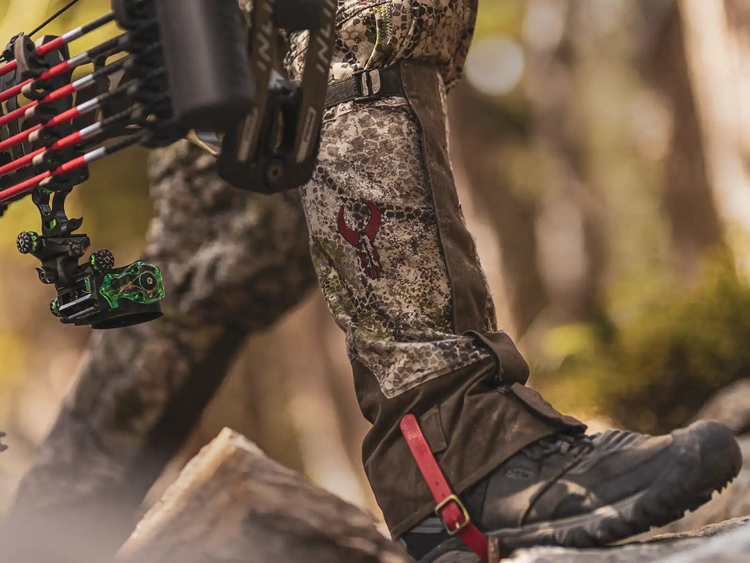 Close-up of camo hunting pants with red Badlands skull logo, black boots, and a bow in a forest setting.