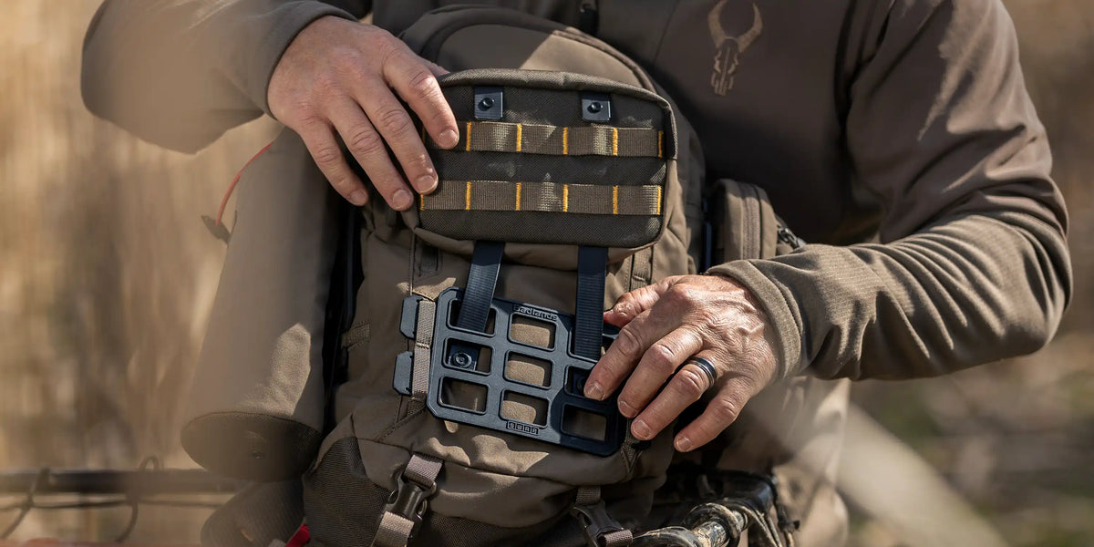 Person attaches gear to a Badlands backpack; Badlands skull logo visible on jacket. Outdoor setting.