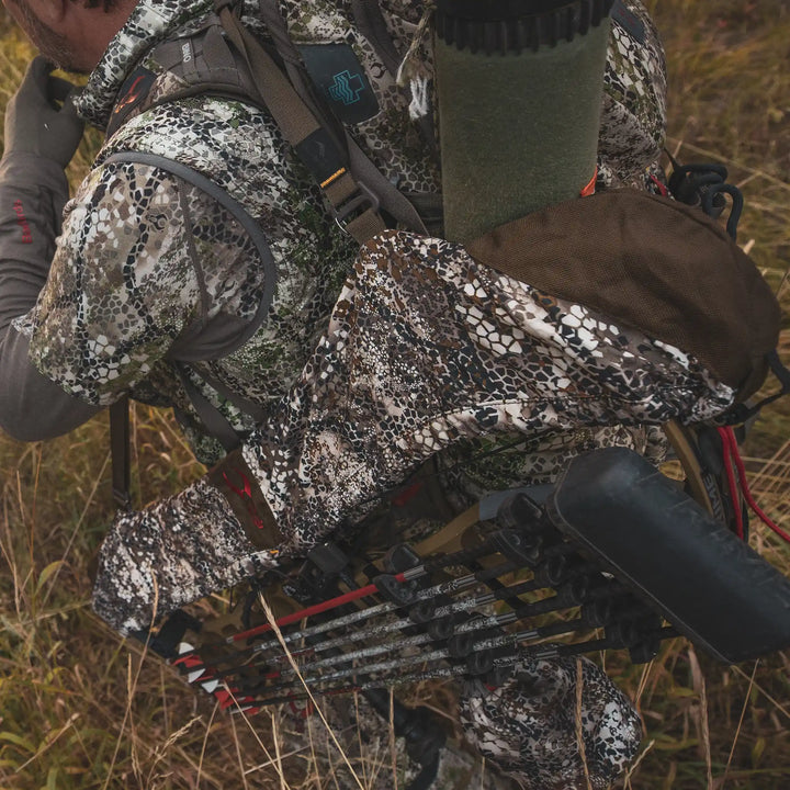 Hunter in camo gear carries a bow in a Badlands pack with the Badlands skull logo, walking through tall grass.