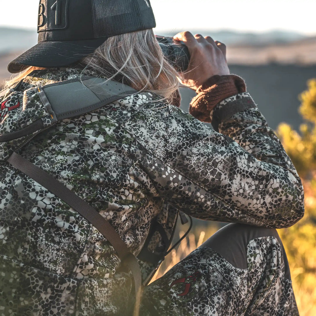 Person in Badlands camo gear with skull logo, using binoculars outdoors; bright, natural setting.