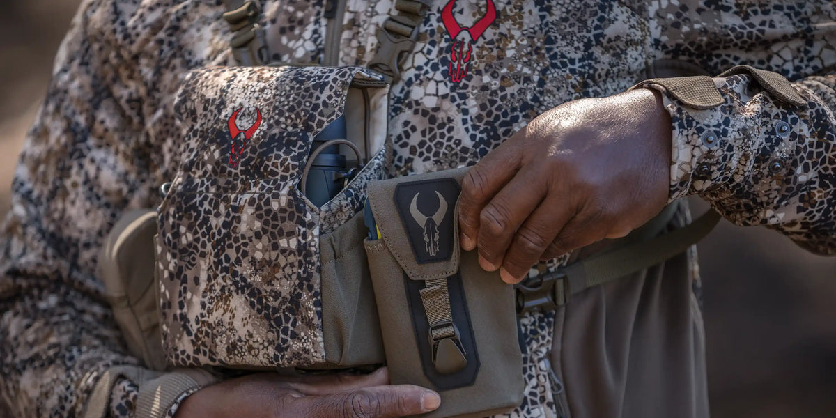 Close-up of hands on Badlands camo gear with red skull logo, showing pouches and textured fabric for hunting or outdoor use.