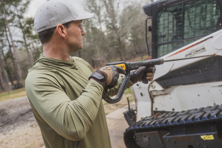 Person wearing Badlands hat and gloves with skull logo uses a pressure washer near heavy equipment outdoors.