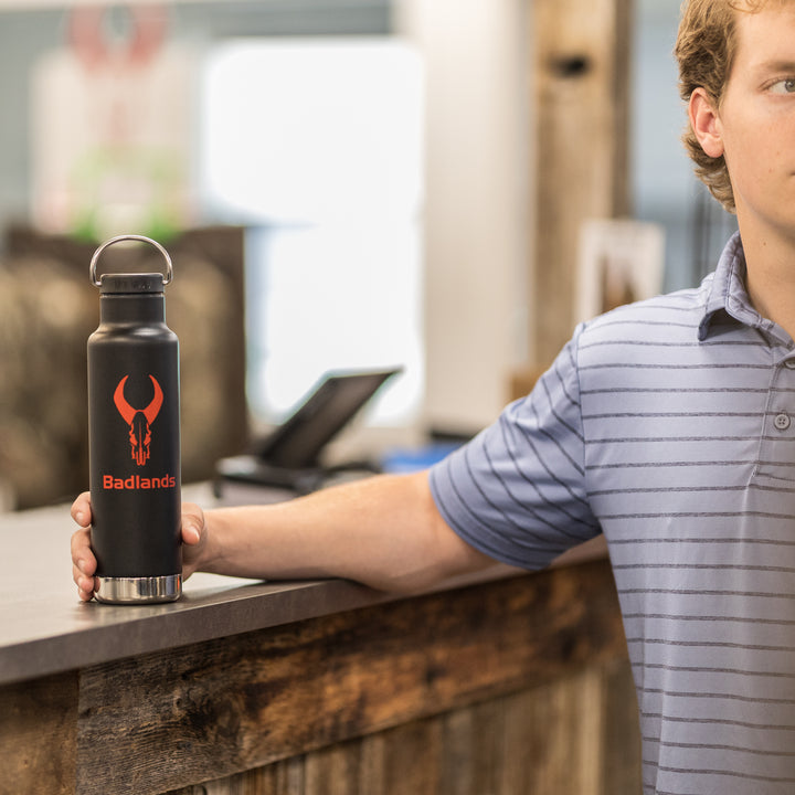 Man in striped shirt holds a black Badlands water bottle with red skull logo on a rustic counter.