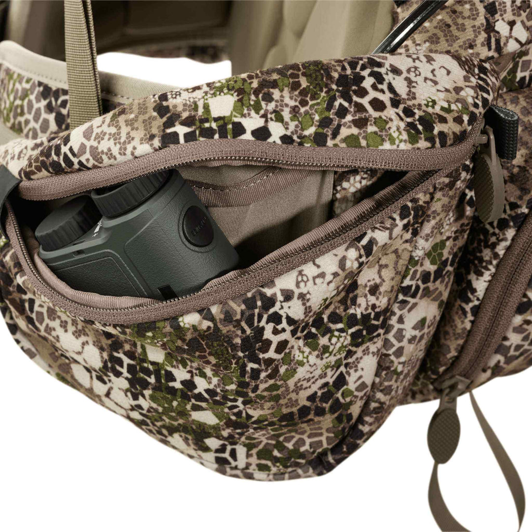 Badlands camo pack with open zippered pocket, showing binoculars inside. Badlands skull logo visible on the fabric.