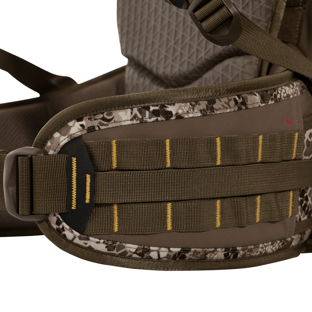 Close-up of Badlands pack hip belt with MOLLE webbing and logo skull patch, shown in camouflage and brown fabric.