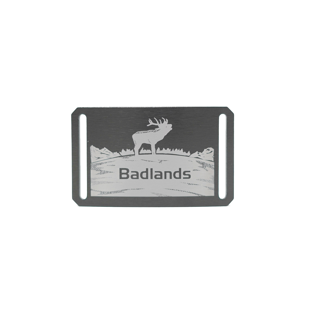 Metal badge with elk silhouette, Badlands logo, and mountain scene; rectangular with two vertical slots for attachment.
