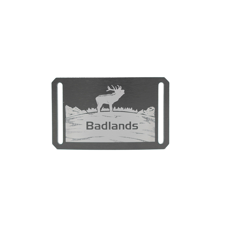 Metal badge with elk silhouette, Badlands logo, and mountain scene; rectangular with two vertical slots for attachment.